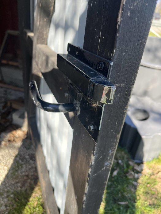 Svart grindlås i metall på trästolpe, soligt, spindelväv, närbild, dagtid, gräs i bakgrund.