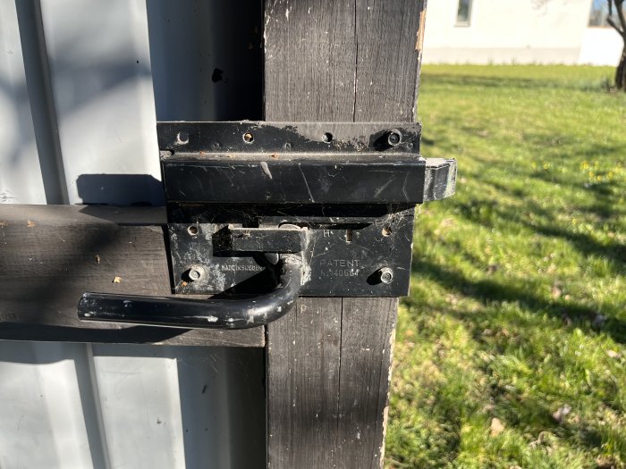 Svart grindlås och handtag på en brun staketstolpe, dagsljus, skuggor, gräs i bakgrunden.