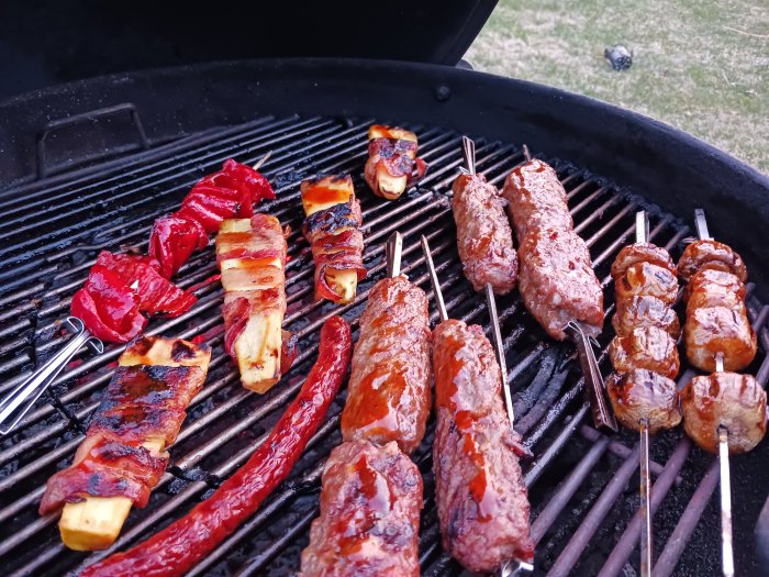 Grill med kebabspett, köttfärsrullar, och grönsaker på glödande grillgaller utomhus.