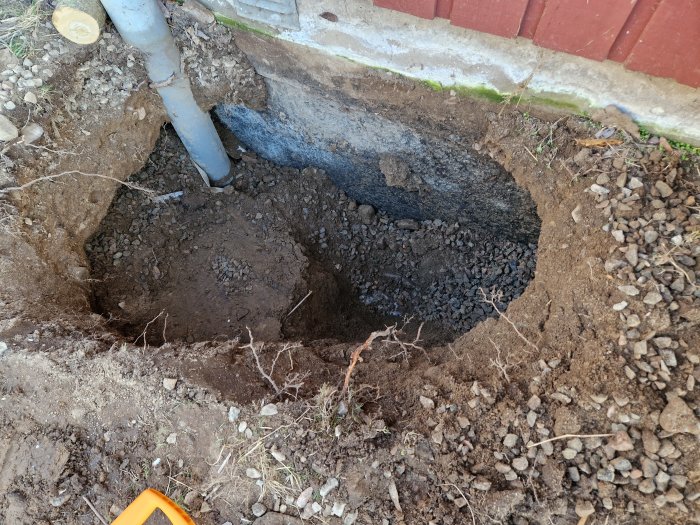 Stort grävt hål i marken bredvid tegelvägg och avloppsrör; grus och jord synlig.