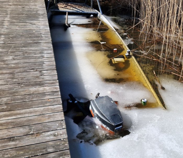 Sunken boat trapped in ice near a wooden dock, surrounded by reeds, under winter sunlight.