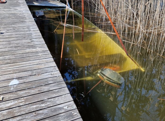 Träbrygga vid vatten, halvsjunken båt, växtlighet, reflektioner i vattnet, soligt, dagtid.