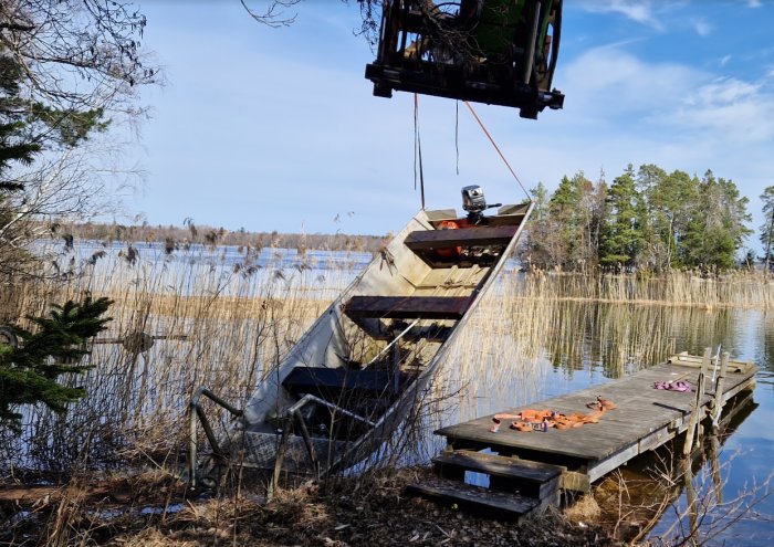 Brygga vid sjö, upphängd båt med motor, verktyg, reed, klar himmel, dagtid, ingen person synlig.