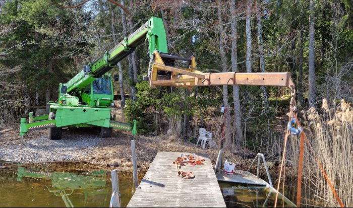 Grön teleskoplastare som använder arm och krok för arbeten vid skog och sjöbrygga.