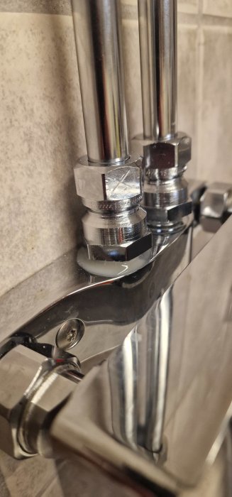 Närbild av kromade rör och anslutningar, troligtvis till en vattenkran, mot kakelvägg.