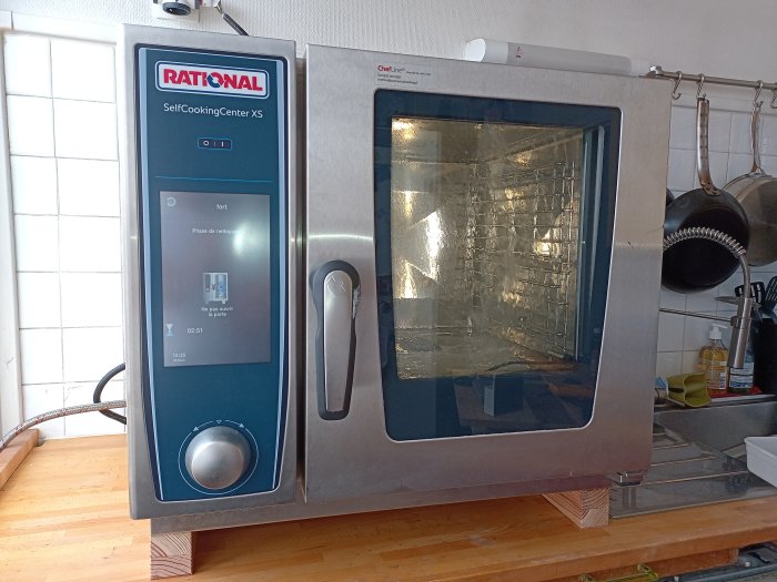 Professionell kombiugn, Rational SelfCookingCenter XS, i köksmiljö, digital display, rengöringsfas, köksredskap i bakgrunden.