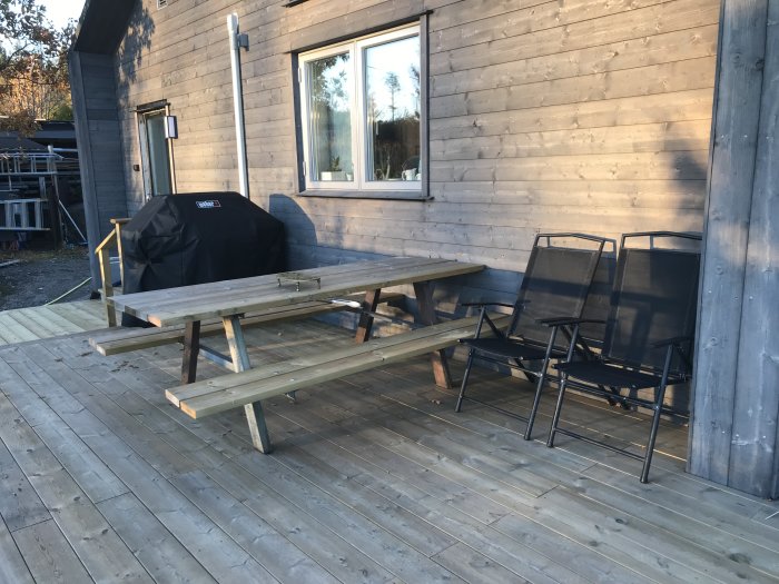 Trädäck med picknickbord, stolar och grill under överdrag vid trähus.