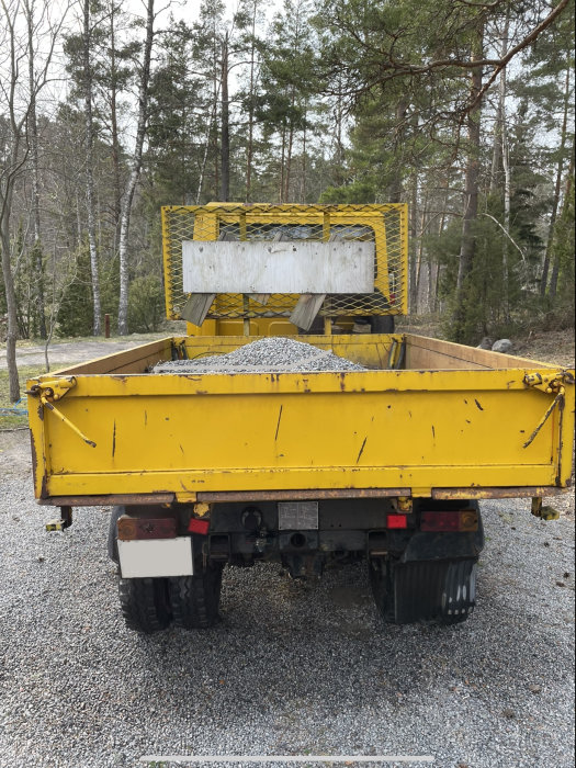 Gul lastbil släpvagn fylld med grus, parkerad i skogsmiljö.