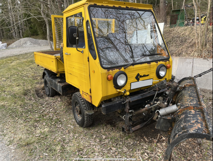 Gul arbetsfordon med flak och snöplog fram, parkerad på grus vid träd.