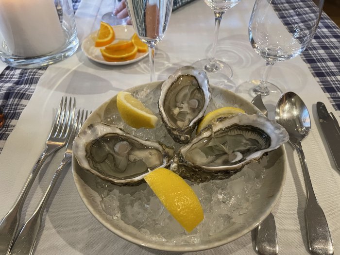 Ett fat med öppnade ostron, citronskivor, is, bestick och glas på bord.