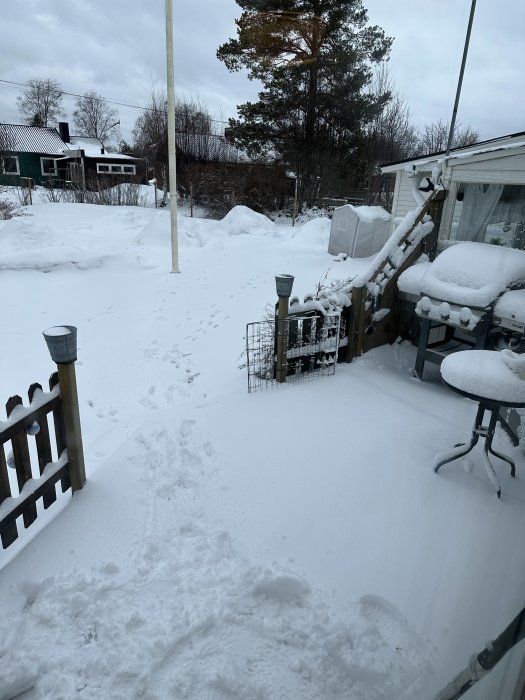 Snötäckt trädgård med möbler och växthus, fotspår på marken, molnig himmel, bostadshus i bakgrunden.