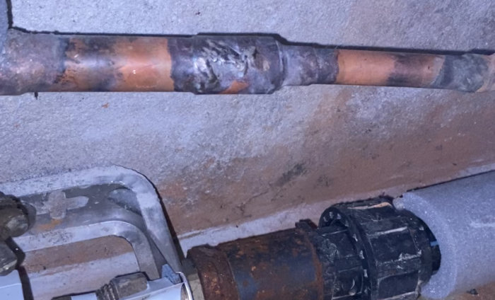 Kopparrör med korrosion och reparationsförsök, vidrörande svart motorpump, på en betongvägg.