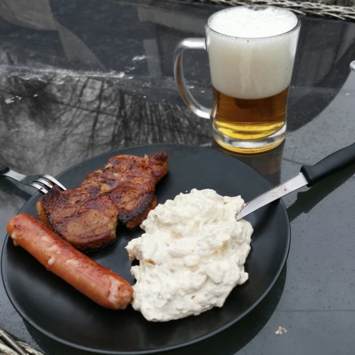 Grillad köttbit och korv med potatissallad på svart tallrik, ett ölglas med skummande öl, utomhusmiljö.