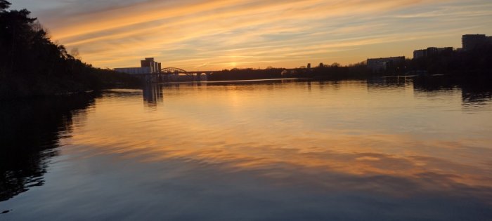 Solnedgång över lugnt vatten med reflektioner, silhuetter av träd och bro, färggrann himmel.