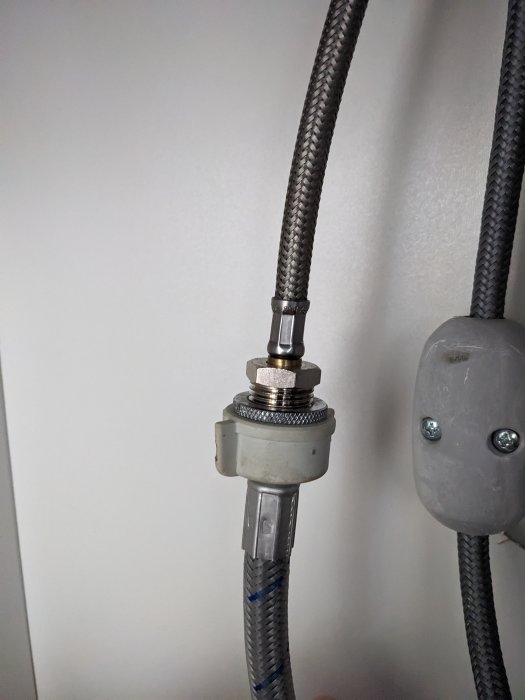 Vattenanslutning med flexibel slang och metallkopplingar mot en vit vägg.