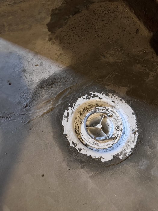 Närbild på en sliten avloppssil i betonggolv med vatten och smuts runtomkring.