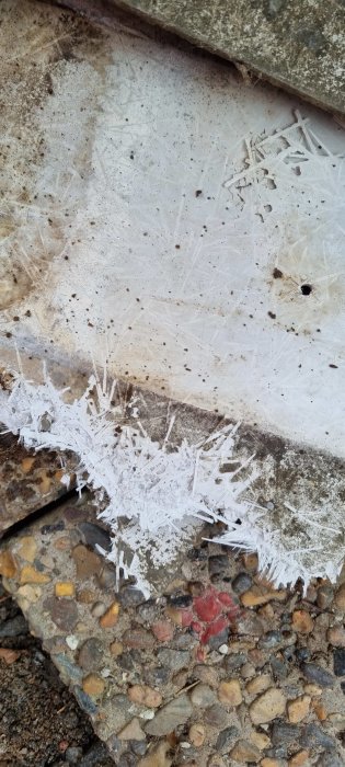 Vertikalt foto av iskristaller som bildats på ojämn betongyta vid grusig markkant.