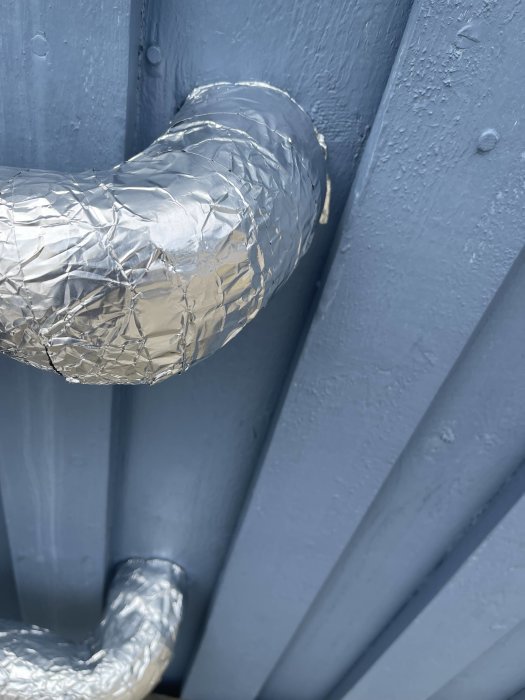 Aluminiumfolie täcker ett rör mot en blå vägg med skruvar och strukturerad yta.