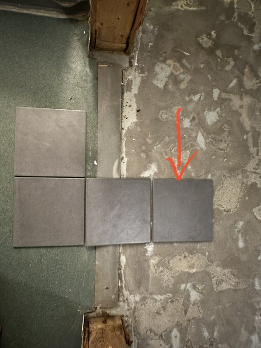 Fyra grå plattor på betonggolv, rött pilmarkering, renovering eller golvinstallation pågår.