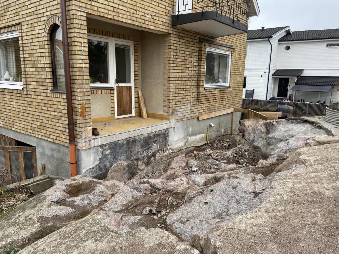 Hus under renovering, utsprängd berggrund, grunden blottlagd, byggarbetsplats, trädäck, dag, molnigt, ingen person synlig.