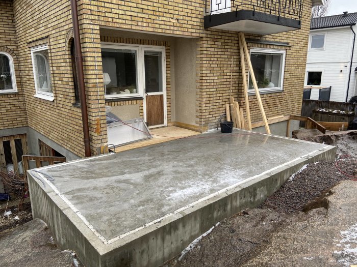 Betongplatta framför hus, delvis täckt av snö, byggmaterial och verktyg synliga, pågående arbete.