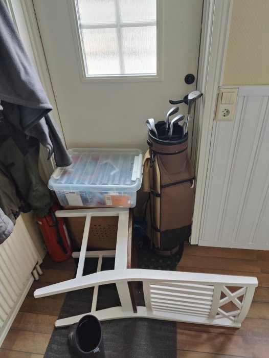 Ingång, golfbag, klubbset, omkullvält stol, förvaringslådor, jacka, svart hink, radiator, fönster och dörr.