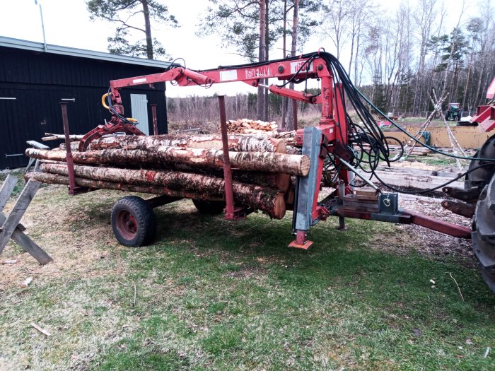 Släpvagn med hydraulisk kran lastad med granstockar, nära skog och byggnad, överkast grått molnigt väder.