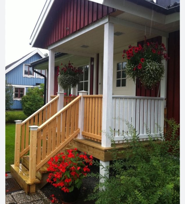 En träveranda med räcke, blommor och en blå husvägg i bakgrunden. Regnigt väder.