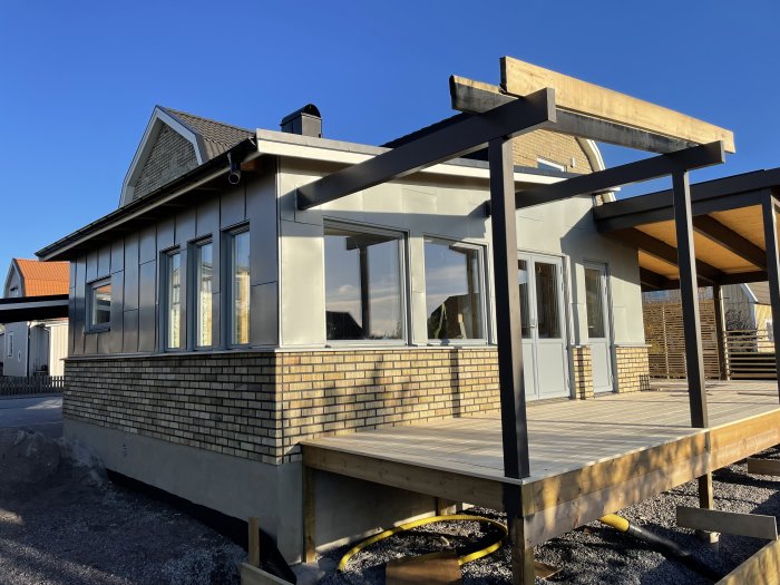 Modernt hus, träterrass, stora fönster, klar himmel, pergola under konstruktion, tegelfasad, bostadsområde, kvällsljus.