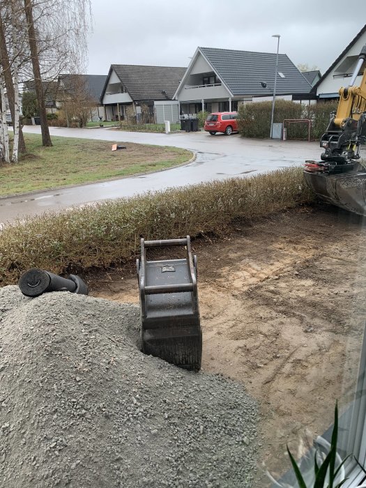 Gråvädersdag, grävmaskin, grushög, tom tomt, bostadshus i bakgrunden, grävd gräsmatta, stillhet, kontrast mellan natur och anläggningsarbete.