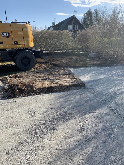 Gul grävmaskin, uppgrävd mark, grusväg, grönska, soligt, del av hus syns, lugnt arbetsområde.