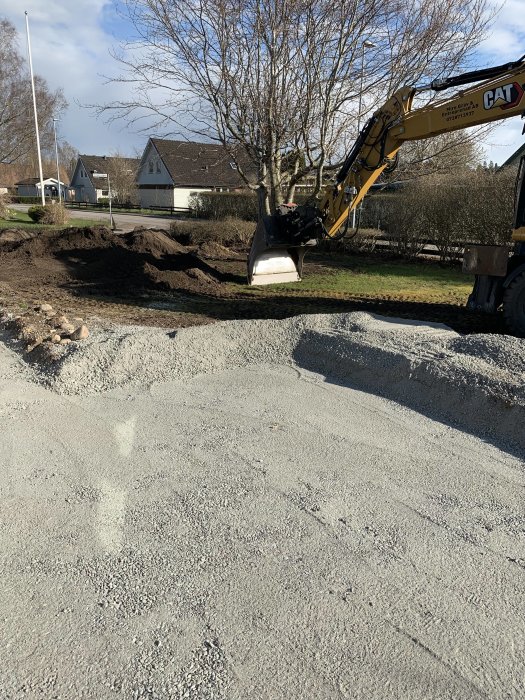 Grävmaskin arbeter vid uppgrävd mark, grus synligt i förgrunden, träd, hus och blå himmel bakgrund.