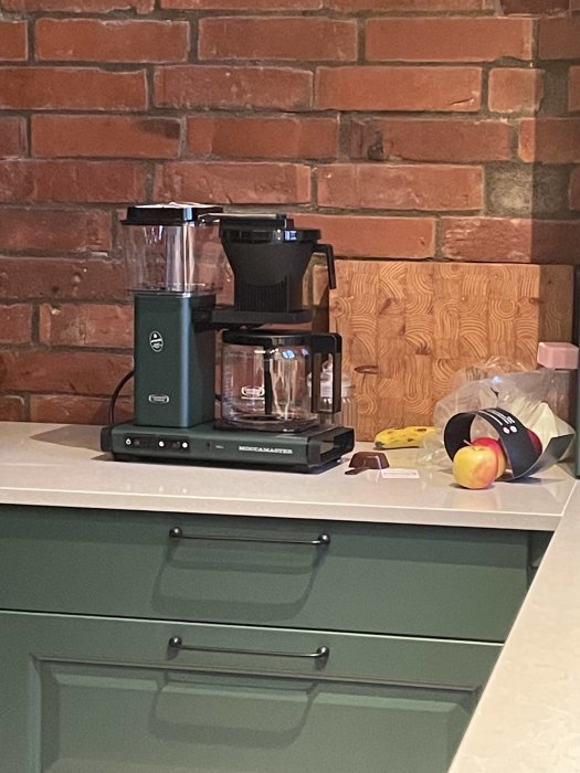 Kaffebryggare på bänkskiva, tegelvägg bakom, skärbräda och frukt framför, modern köksinredning.