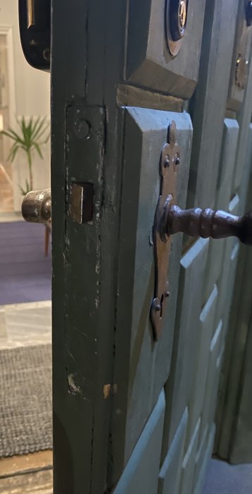 Grönmålad trädörr med traditionella dörrhandtag och lås, tecken på slitage, inomhusmiljö syns i bakgrunden.