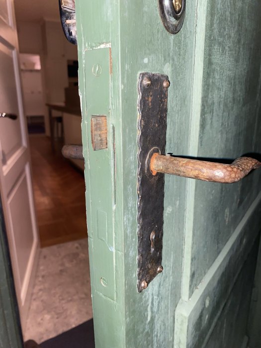 Grön dörr på glänt med rostig, gammal handtag och låsmekanism. Inredning synlig i bakgrunden.