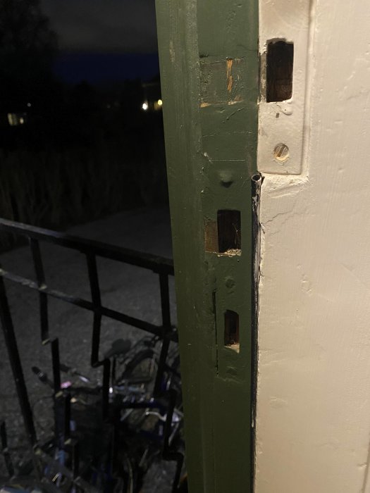 Grön dörrkarm med luckor där gångjärnen varit. Vit vägg. Kväll. Cykelställ i bakgrunden.