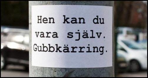 Lapp med text "Hen kan du vara själv. Gubbkärring." klistrad på stolpe, bakgrund med oskarpa fordon.