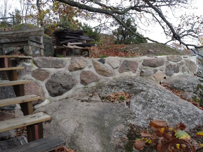 Stentrappa, stenmur, träd, höstlöv, staplade brädor, naturmiljö, grått klippblock, utomhus, dagsljus, ingen himmel synlig.