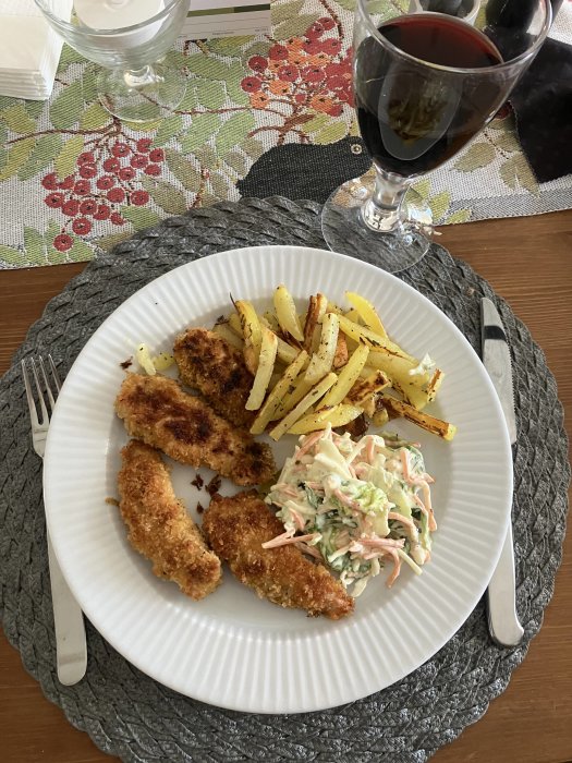 Måltid med panerad kyckling, pommes, coleslaw, rödvin, vattenglas, bestick, blommig bordstablett.