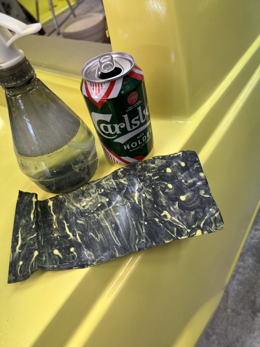 En öppnad Carlsberg ölburk, en genomskinlig sprayflaska, och en bit plastfolie på ett gult bord.