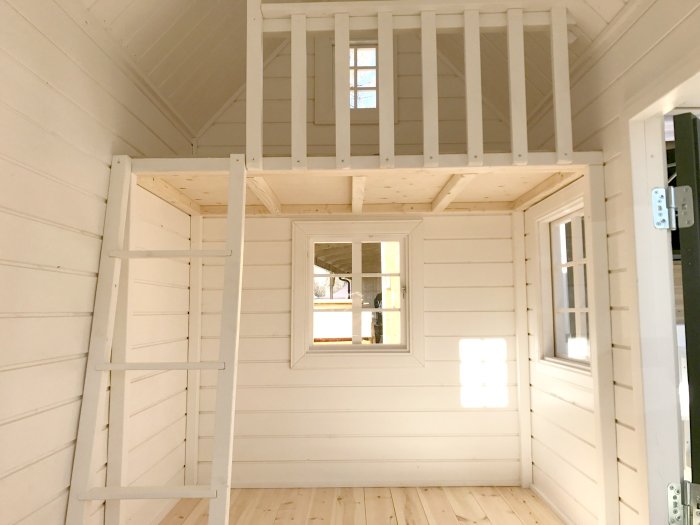 Ljust trälekstuga interiör, liten lofttrappa, fönster, öppen dörr, vitmålat, enfärgat, minimalistiskt.