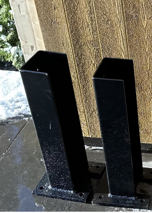 Två svarta metallstolpar monterade vid en trävägg, med snö och is i bakgrunden.