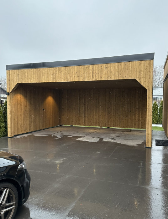 Modern träbeklädd carport, regnvåt uppfart, del av svart bil, grå himmel, gräsmatta.