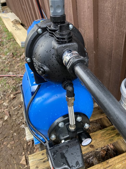 Blå cistern med pump och rör, fuktig yta, monterad utomhus vid trästaket.