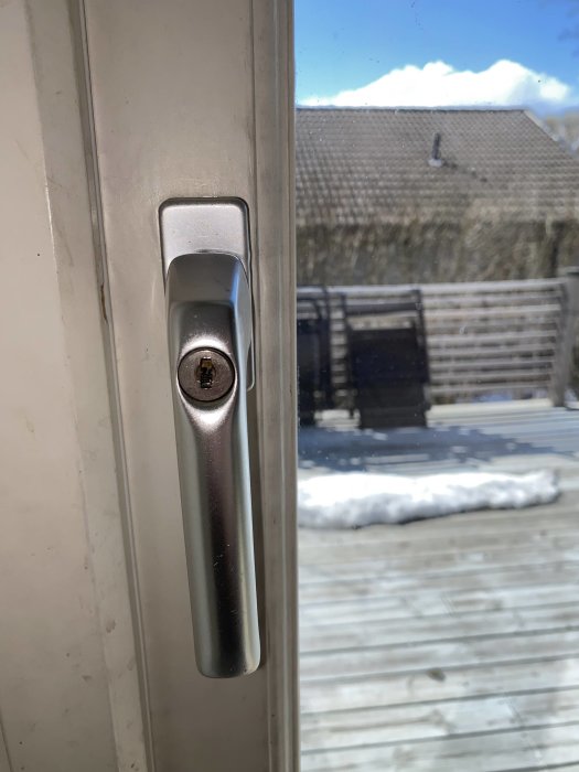 Fönsterhandtag inifrån, utsikt över trädäck med snö, soldis, tak i bakgrunden.