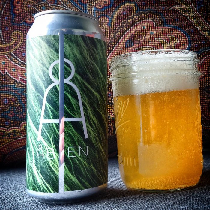 Ölburk med logotyp bredvid ett glas skummande öl mot mönstrad bakgrund.