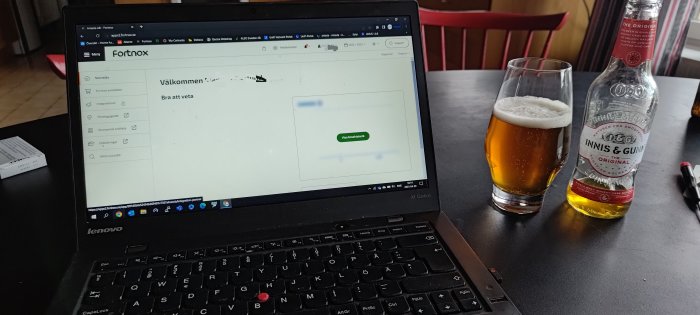 Laptop med öppen webbsida, ölglas och tom ölflaska, på bord inomhus.