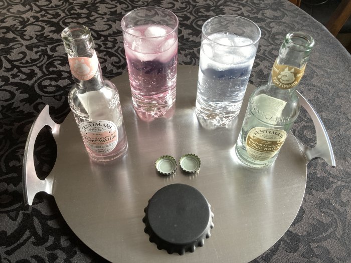 Två flaskor tonicvatten, färgade glas med is, kapsyler, palettformad bricka, mönstrad yta.