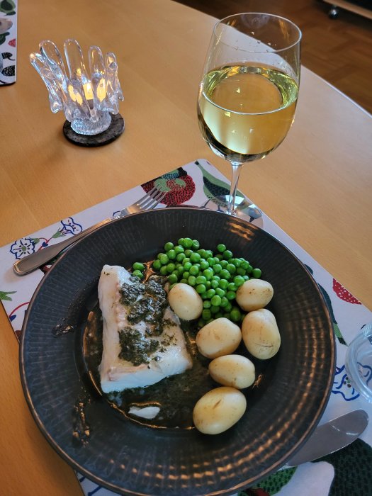 Fisk med sås, gröna ärtor, kokta potatisar och ett glas vitt vin, stämningsfull belysning i bakgrunden.