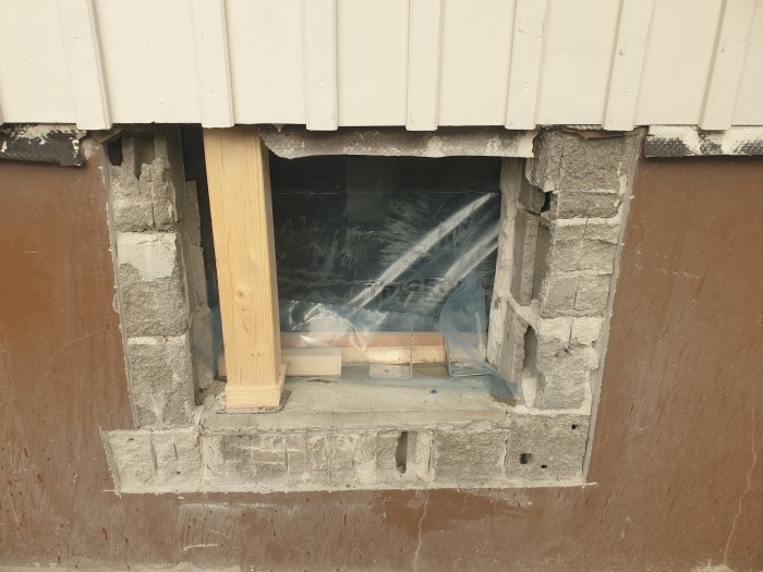 Fönsteröppning under reparation, plastskydd, trästöd, avskalad fasad, ouppklart byggprojekt, murblock, exteriör vägg.